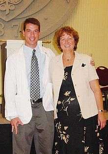 The same man seen in the infobox, here shown smiling with his teeth showing wearing a white doctor's coat over a shirt and tie, standing on the left of a smiling older woman with red hair and rosy cheeks wearing a black print dress with a pale yellow jacket. Behind them is a tage with a portion of a large seal visible