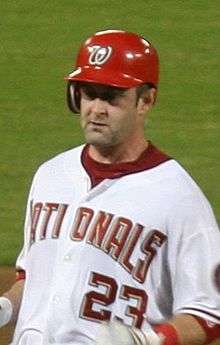 A man wearing a white baseball jersey and red batting helmet with a look of concentration upon his face