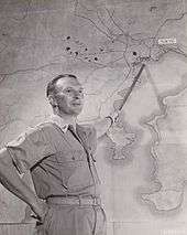 Black and white photo of a middle-aged man wearing military uniform pointing at a map of the Toyko Bay region of Japan with a stick