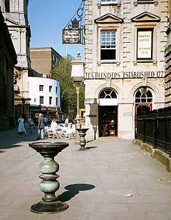 Corn Street, Bristol. A stone-paved pedestrianised street surrounded by classical architecture. In the foreground are two ornate cast bronze tables known as a nails.