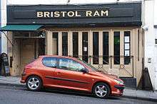 Photo of a building with slatted windows, and awning above door. An overhead sign reads Bristol Ram. A red car is parked in front on a road sloping down to the right