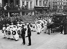 A procession of Suffragettes, dressed in white and bearing wreaths and a banner reading "Fight on and God will give the victory" during the funeral procession of Emily Davison in Morpeth, Northumberland, 13 June 1913. Crowds line the street to watch.