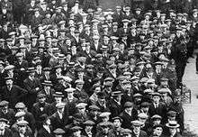 A large throng of people congregate, surrounded by police officers. Approximately half wear peaked caps; the rest wear boaters. Some smile, the rest look pensive.