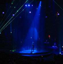 Faraway image of a female performer. She is walking through a water screen that is falling from the ceiling. A blue spotlight is on her. She is wearing blue jeans and a plastic cowboy hat. Green laser lights surround the stage.