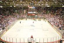 The view of an ice hockey rink from behind and above one goal. Most players, dressed in either red and white or blue and black, are in the far right corner of the rink. Above center ice an electronic scoreboard reads "9:09" in green; under that, red numbers read from left to right "0", "1", and "0". The "1" is smaller than the two "0"s. On the visible end of the rink, a red sign reads in black and white text "Pizza Hut".