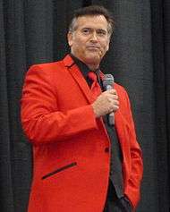 A man standing with a microphone, wearing a red blazer.