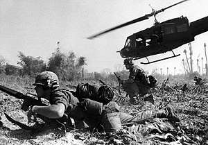 A black and white image of a man crawling across the ground with a helicopter flying in the background