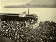 A dramatic political scene. Beside a river stands a podium, on which a flagpole flies a huge American flag. Beneath the flag stands a candidate in a dark suit addressing an impressive crowd that takes up most of the photograph. Not only the quayside, but a ferry beside it on the water are packed full of people listening intently.