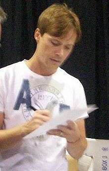 A fair haired young man wearing a white T-shirt, signing a piece of paper