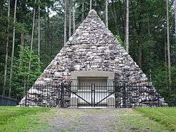 A fieldstone pyramid surrounded by a fence with pine trees in the background