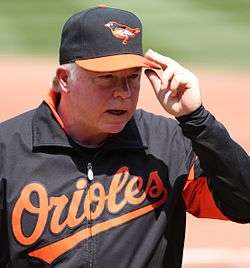 A man in a black jacket with "Orioles" written across the chest in orange script letters and a black and orange cap with an oriole stitched on it touching the bill of this cap