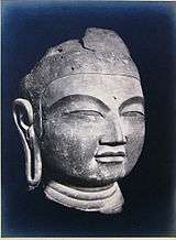 A Buddha head in three-quarter view with elongated earlobe, thick lips, narrow slit eyes and broad nose.