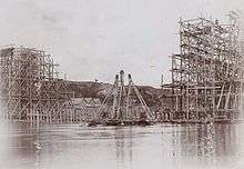 Sepia photo showing construction of a bridge across the Neman. On both sides completed parts of the bridge surrounded by wooden scaffolding are visible. In between a wooden pyramid-shaped floating crane is being assembled.