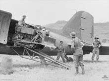A tiny bulldozer emerges from the rear door of an aircraft onto a grass airstrip. Three men stand around watching.