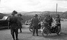 black and white photograph of a military car and two motorcycles with dismounted soldiers, some with a plume of feathers attached to their helmets
