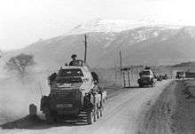 An eight-wheeled armoured car on a dirt road with other vehicles in the background