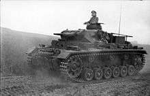 a black and white photograph of a tank driving along a dusty road