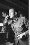 Black-and-white portrait of a man on the phone in semi profile wearing a military uniform with an Iron Cross displayed at his neck. He is holding a cigarette in his left hand.