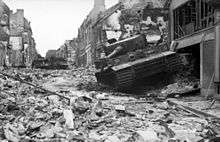 Three stationary tanks, one in the foreground and two in the rear, in a heavily damaged street; rubble covers the road.