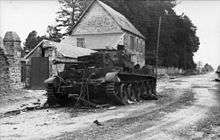 A knocked out tank sits at the side of a road, in front of a two-story house.