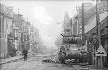 A soldier walks up a high street; the sides of the road are covered with rubble. In the foreground there is a knocked out tank, at the foot of the tank there is a dummy wooden gun barrel and debris. Another knocked out tank being examined by another solder occupies the background.