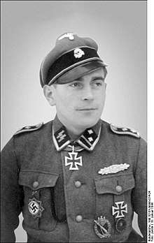 A man wearing a peaked cap with skull emblem, a military uniform with various military decorations and an Iron Cross displayed at the front of his uniform collar.