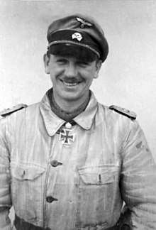 A black-and-white photograph of a smiling man wearing a military uniform, peaked cap and neck order, in shape of an Iron Cross.