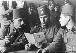 three soldiers in SS uniform and wearing fez headgear reading a pamphlet