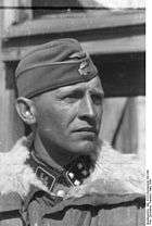 A black-and-white photograph of a man wearing a military uniform with fur collar, side cap and a neck order in shape of an Iron Cross. His cap has an emblem in shape of a human skull and crossed bones.