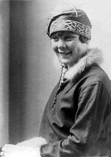 A young smiling woman wearing an embroidered hat and a jacket with furred collar and sleeve hems.