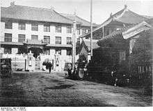 A black and white image of several buildings with Chinese-style wide-eaved roofs making a corner around a parking area where a 1920s-style automobile is parked