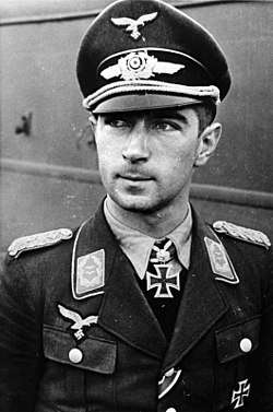 The head and shoulders of a young man. He wears a uniform and peaked cap, with an Iron Cross displayed at the front of his shirt collar.