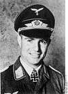 A smiling man wearing a military uniform, peaked cap, and an Iron Cross displayed at the front of his uniform collar.