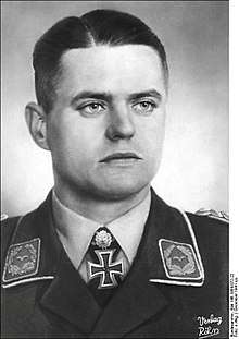 The head and shoulders of a young man, shown from the front. He wears a military uniform, with an Iron Cross displayed at the front of his white shirt collar. His facial expression is a determined and a grim smile; his eyes are looking to the left of the camera.