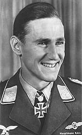 The head of a young man, shown in semi-profile. He wears a military uniform with a military decoration in shape of an iron cross displayed at the front of his shirt collar. His hair is dark and short and combed to back, his nose is long and straight, he is smiling broadly and looking to the left of the camera.