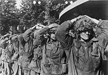 Eight men walking towards the camera with the hands on their heads