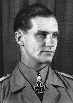 The head and shoulders of a young man, shown in semi-profile. He wears a military uniform with an Iron Cross displayed at the front of his shirt collar. His hair appears blond and short and combed back, his nose is long and straight, and his facial expression is determined but smiling; looking to the left of the camera.