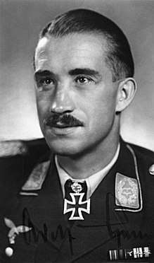 The head and shoulders of a young man, shown in semi-profile. He wears a military uniform with various medal ribbons above his left breast pocket and an Iron Cross displayed at the front of his shirt collar. On his upper lip is a moustache, his hair is dark and short and combed back, his facial expression is a determined and confident smile; his eyes gaze into the distance.
