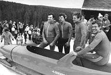 Four men in winter tracksuits stand at the side of a bobsled. They are outdoors, at a snow-covered hill top, with many people behind them.
