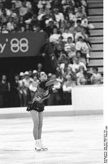 A female figure skater points with her right arm as she performs