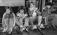 Four women wearing tracksuits and speed skates are sitting on a bench. Three of them are smiling, and among these, two are tying up their skates while a third one is clapping her hands.