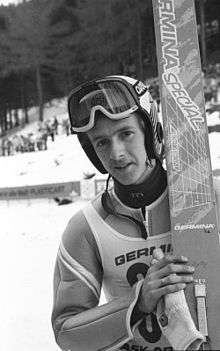 A man is shown with a white vest bearing the word Germany and a number printed in black on top of a ski jumping suit. He also wears a ski helmet with attached goggles and holds his skis upright over his left shoulder.