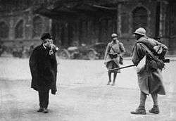 A soldier, on the right, faces a civilian, on the left. A second soldier, far center, walks towards the two.