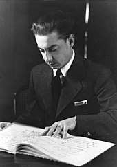 young clean-shaven man, with dark, slicked back hair; he is studying a musical score