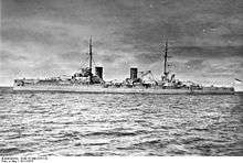 A large gray warship with four large gun turrets and two tall funnels sits idly in harbor.