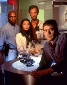 Four young, scientist characters—one Asiatic woman, two Caucasian men, and one African-American man—standing in a laboratory-like set. They look at the camera with glum expressions.