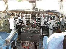 DC-7 cockpit viewed inside, facing forward. Three vertical pillars support a windscreen with two panes. Forty round instrument dials are on a flat panel in front of two seats. Facing each seat is a short column topped with a semi-circular control yoke. Between the seats are two clusters of engine throttles, one set of four for each pilot.