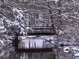 Snow covers a simple wooden footbridge which crosses a stream in a forest