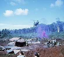 Colour image of a large two-rotored helicopter flying past a line of trees and preparing to land in an open area cleared of vegetation. In the foreground are a large number of tents, hootchies and ground sheets, while a number of soldiers are standing around an artillery piece. Purple smoke is rising from the position.