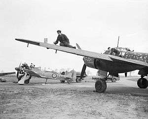 A photograph of a single–engined and a larger, twin engined aircraft on the ground. Two men are working on the wing of the larger aircraft. The engine–covers have been removed from the smaller aircraft and four men are working on its engine